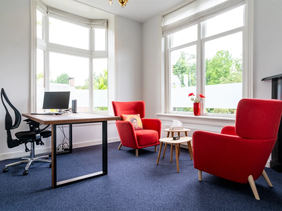 afkickkliniek-fazantlaan-behandelruimte-rode-stoelen Lichte therapieruimte, met een werkplek voor de behandelaar en 2 rode fauteuils tegenover elkaar.