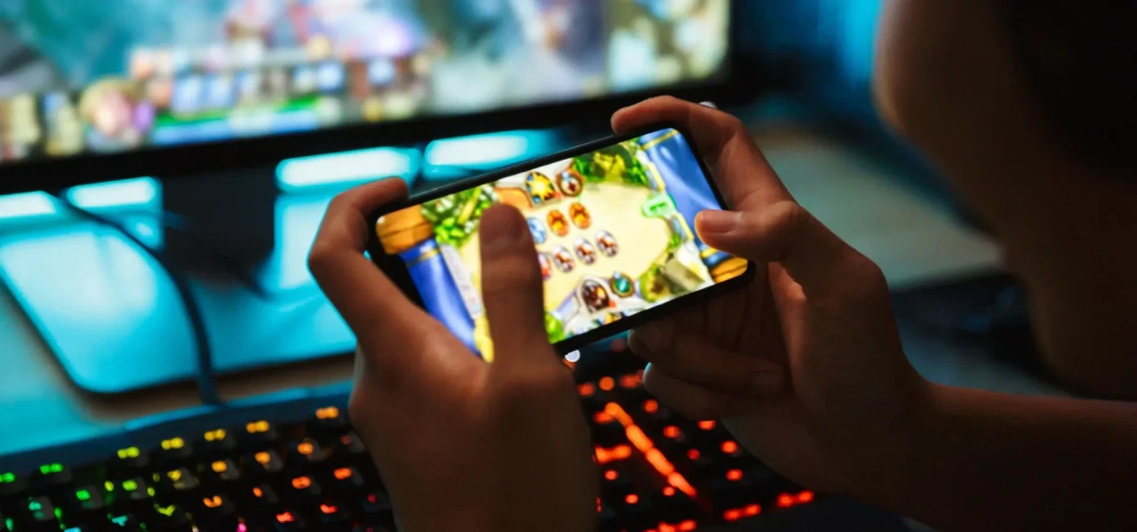 Iemand met een gameverslaving houdt met twee handen een telefoon vast en speelt een spelletje op zowel de telefoon als computer. 