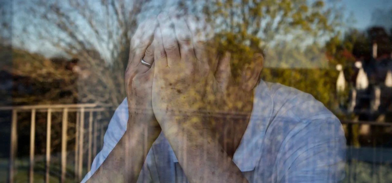 Depressieve man met hoofd in de handen, zichtbaar door semi-weerspiegelend raam.