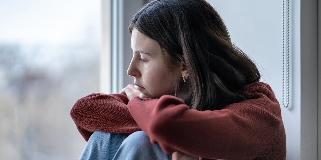 Vrouw met burn-out zit bij raam en staart somber naar buiten.