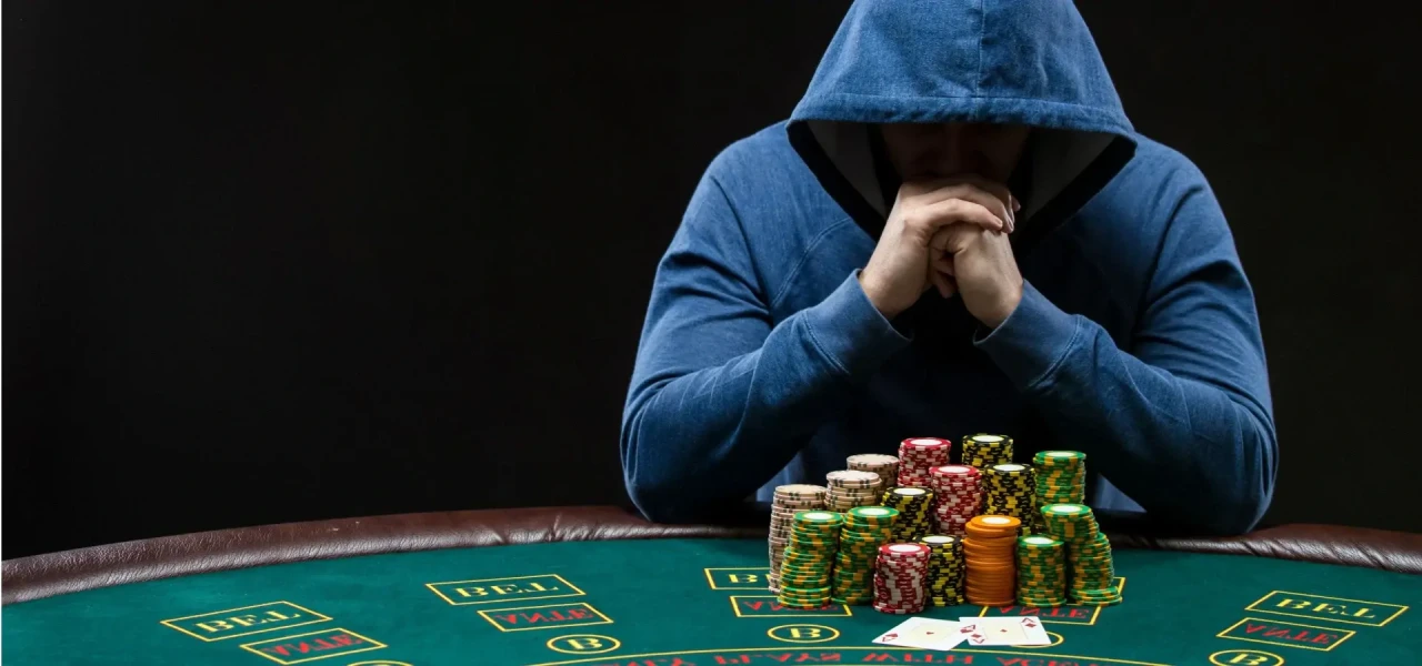 Man in blauw vest met pokerverslaving zit achter een groene pokertafel met fiches voor zich.