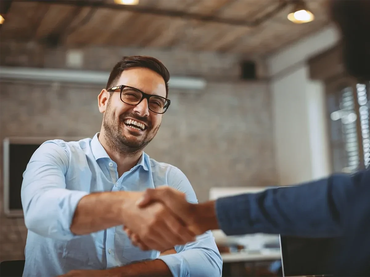 lachende-man Lachende man in blauw overhemd en bril die vacature afneemt en de andere persoon de hand schudt.