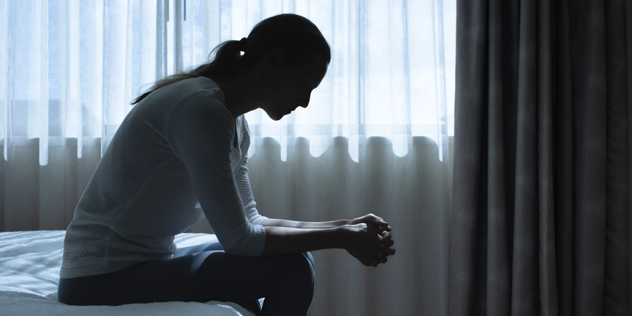 Vrouw zit voorovergebogen op bed bij raam, silhouet dat PTSS uitbeeldt.