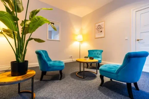 Behandelkamer met drie blauwe stoelen om een kleine salontafel heen. Daarnaast staat een stalamp en een tafeltje met groene plant.