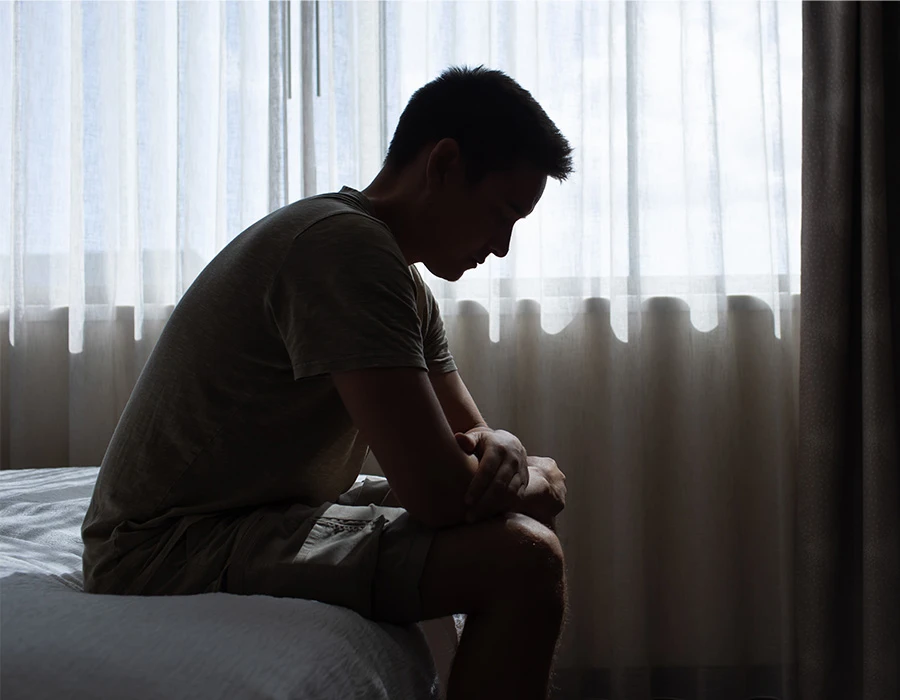 Man zit alleen op bed met gebogen houding, zichtbaar somber.