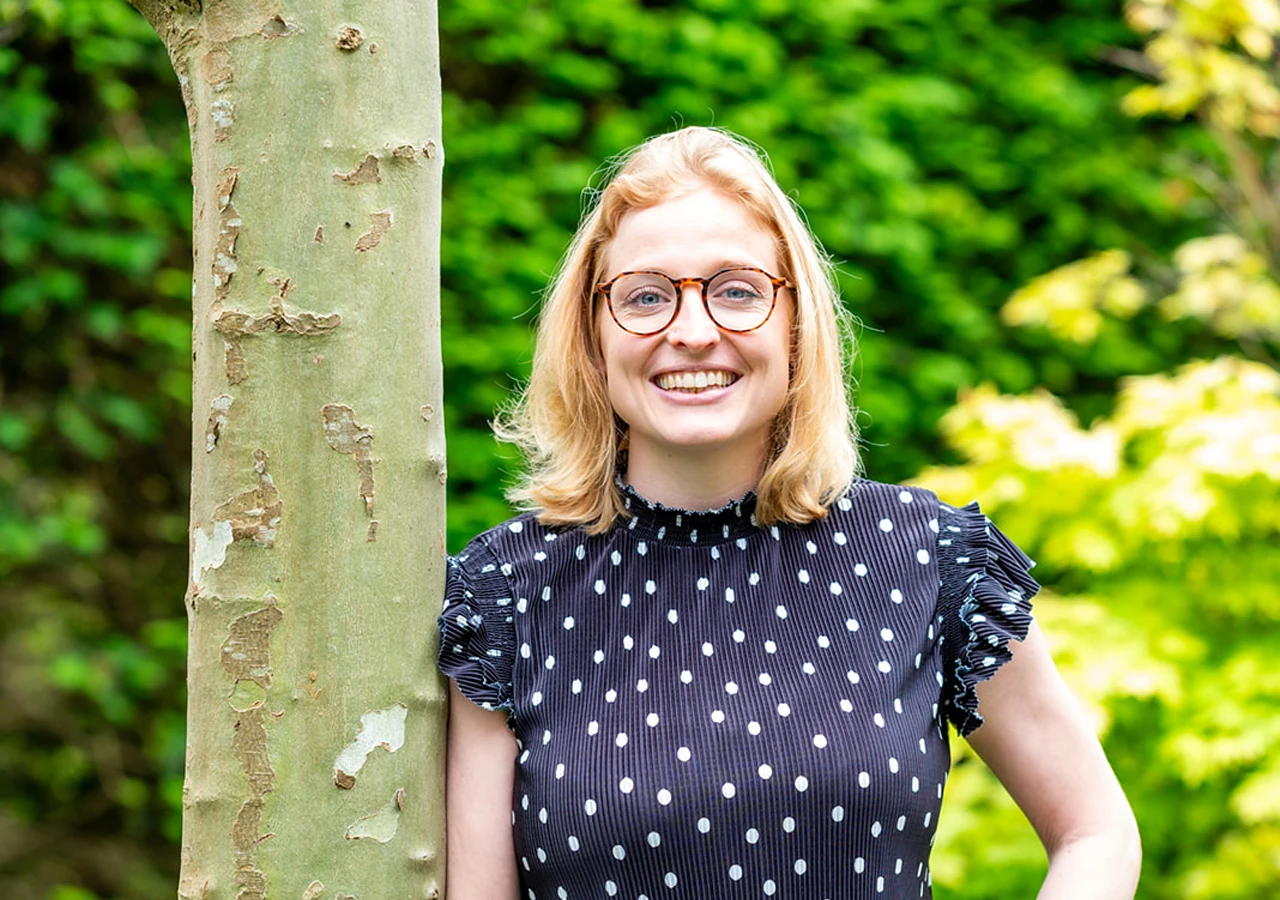 Lachende vrouw met blond haar, een bril en een blauw shirt met stippen staat tegen een boom geleund met een groene achtergrond van wazige bomen en planten.