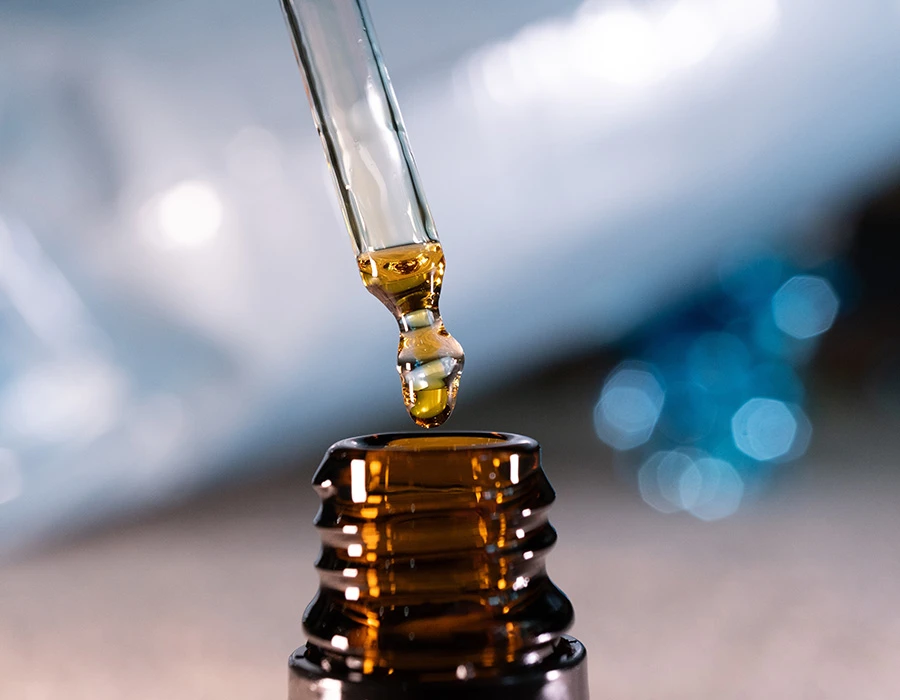 Druppel olie aan een pipet boven een bruin flesje, ter illustratie van CBD-olie.
