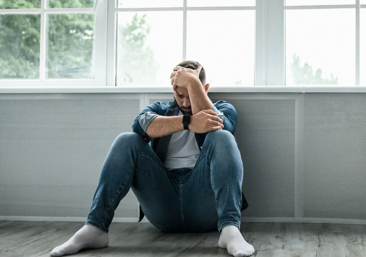 depressieve-man-zittend-tegen-raam Depressieve man met spijkerbroek en witte sokken zittend tegen een raam met hand op het hoofd.