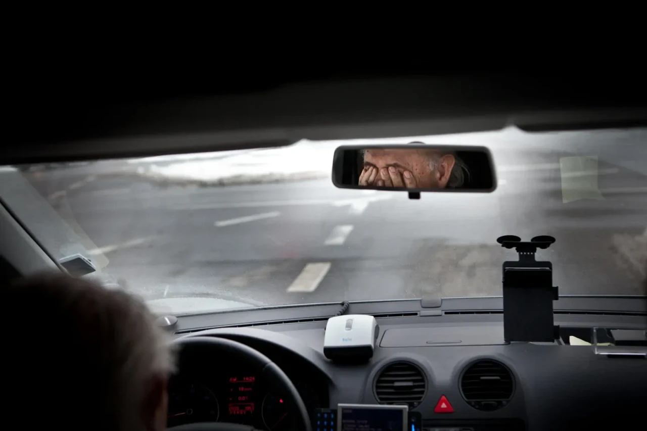 Binnenkant van auto, waarin je in de achteruitkijkspiegel een depressieve man zijn handen voor de ogen ziet slaan.
