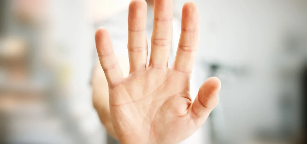 Close-up van een hand die gebaart te stoppen, tegen een onscherpe achtergrond van een man in wit T-shirt.