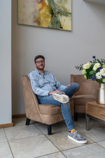Wesley, met pet, bril en baardje, vertelt zijn herstelverhaal terwijl hij in een bruine stoel zit.