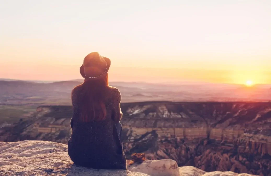 vrouw-kijkt-naar-zonsondergang Vrouw zit op een rots en kijkt naar de zonsondergang over een natuurgebied uit.