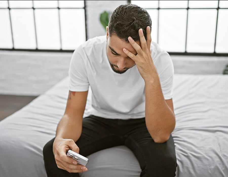 Man met ADD kijkt bezorgd naar telefoon terwijl hij op bed zit.