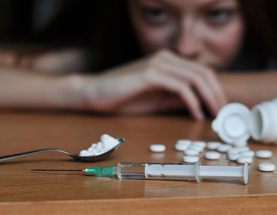 Spuit met drugs en pillen op tafel met persoon op achtergrond.