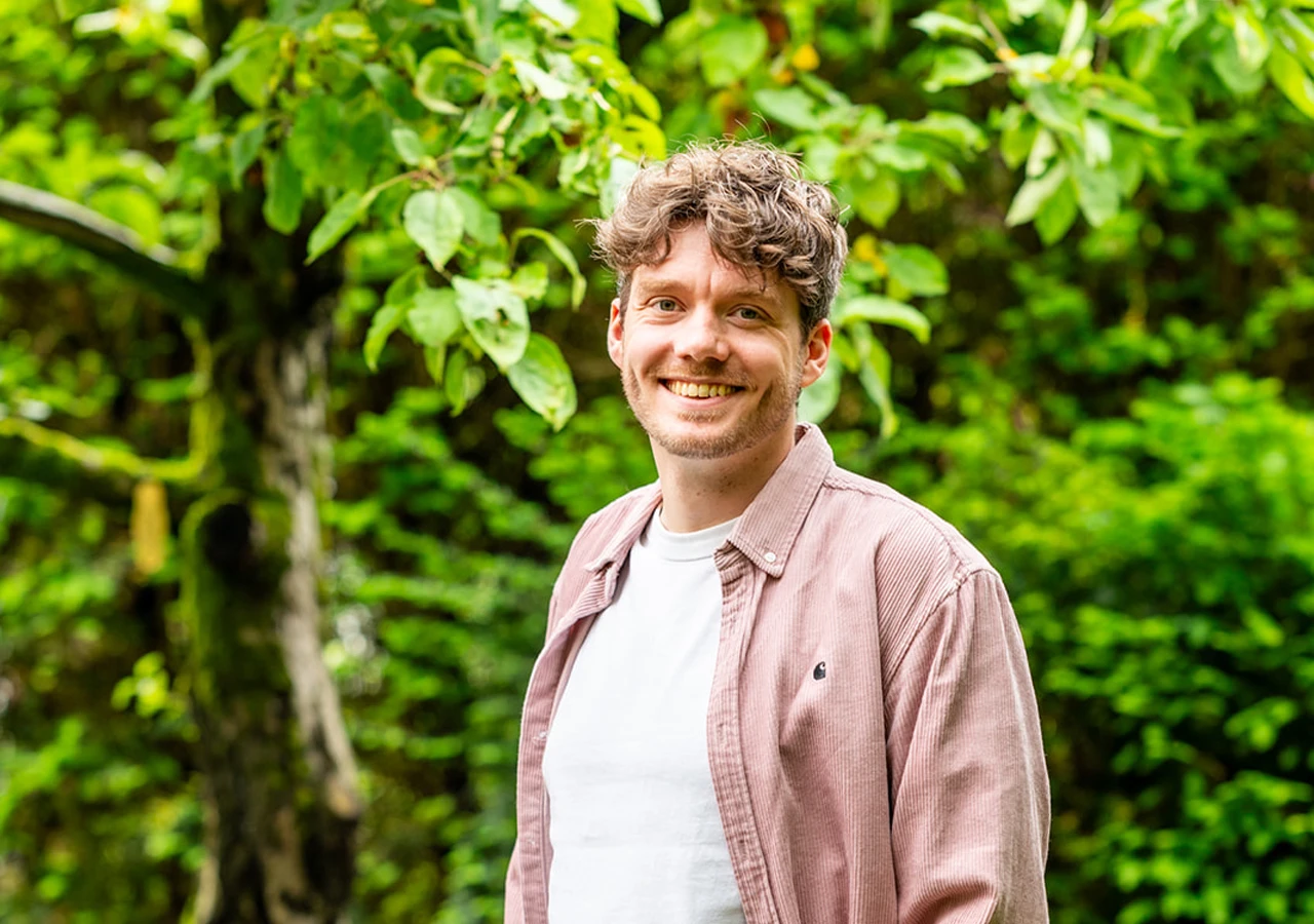 Lachende man met een wit shirt en roze blouse staat voor een groene achtergrond van wazige bomen en planten.