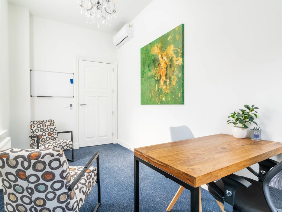 Behandelruimte met witte muren en een groen schilderij, blauw tapijt, een houten bureau en twee stoelen met drukke print.