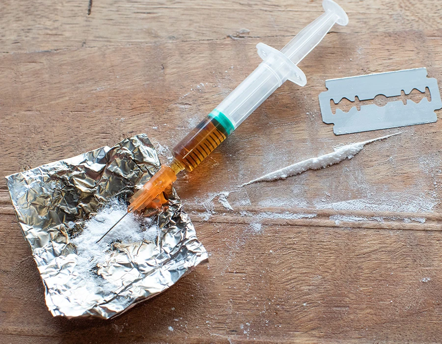 Close-up van een injectiespuit naast wit poeder en folie op tafel, passend bij harddrugsgebruik en crackverslaving.