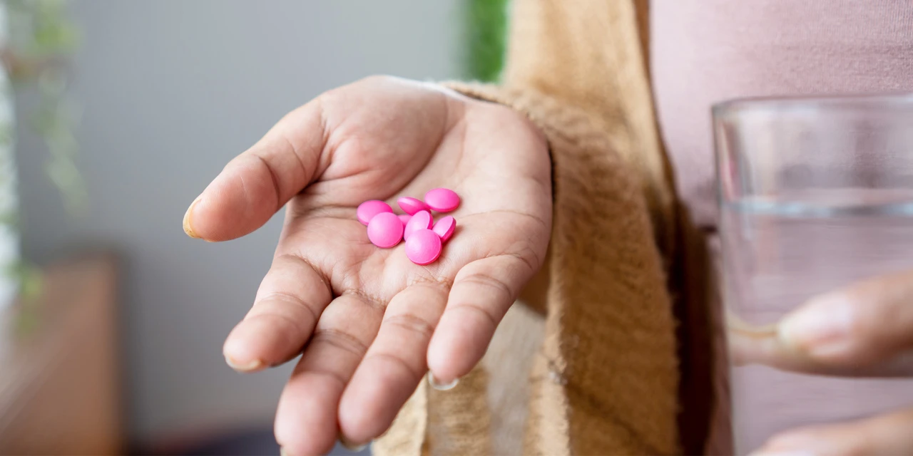 Hand met roze pillen naast glas water waarschuwt voor ibuprofen- en alcoholgebruik.