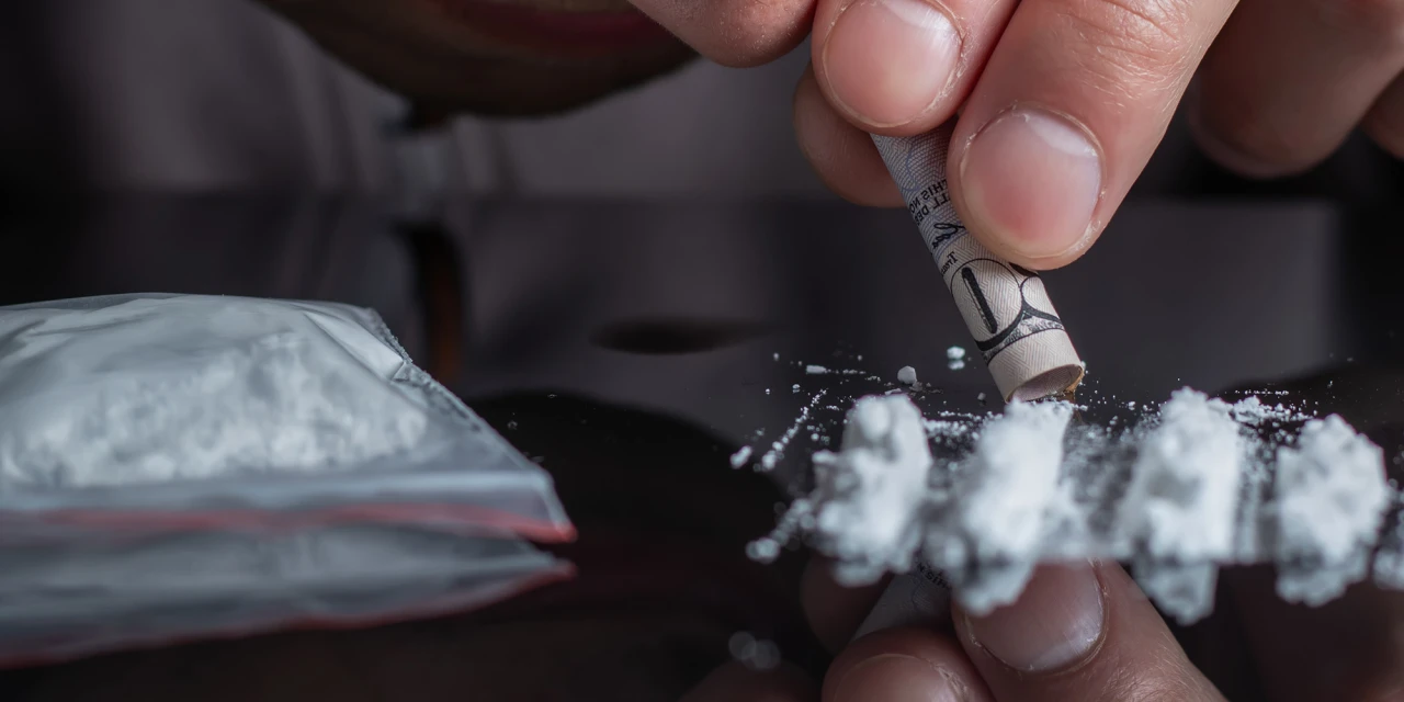 Close-up van persoon die coke snuift met opgerold biljet naast hoopjes cocaïne.