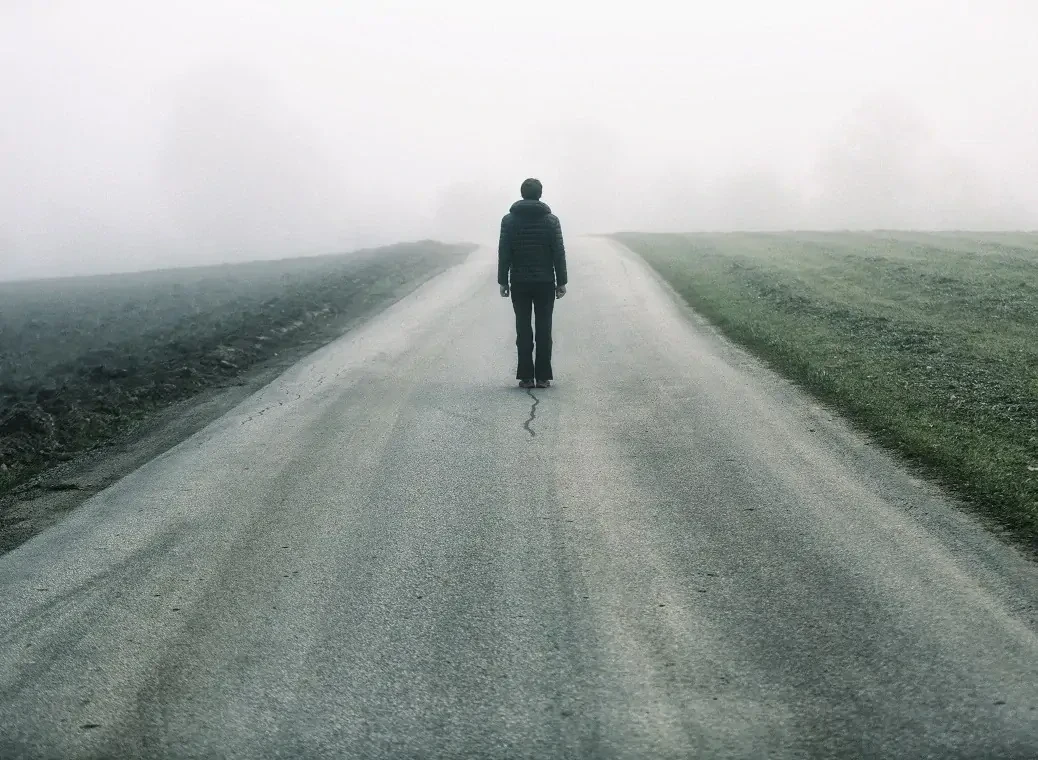eenzame-man-op-weg Man loopt op een lege, mistige weg, met aan weerskanten grasvelden.