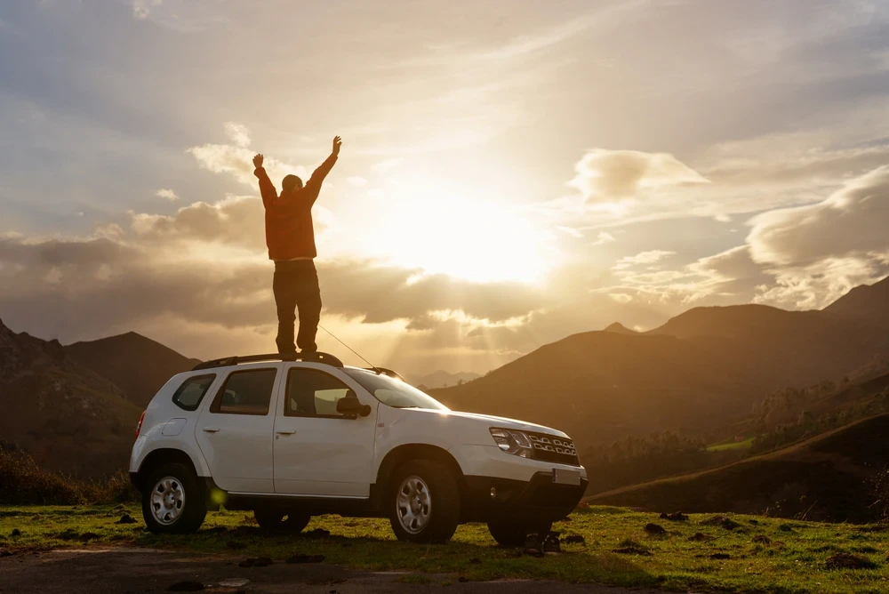 Man staat op witte auto in natuurgebied met de zon en bergen op de achtergrond.