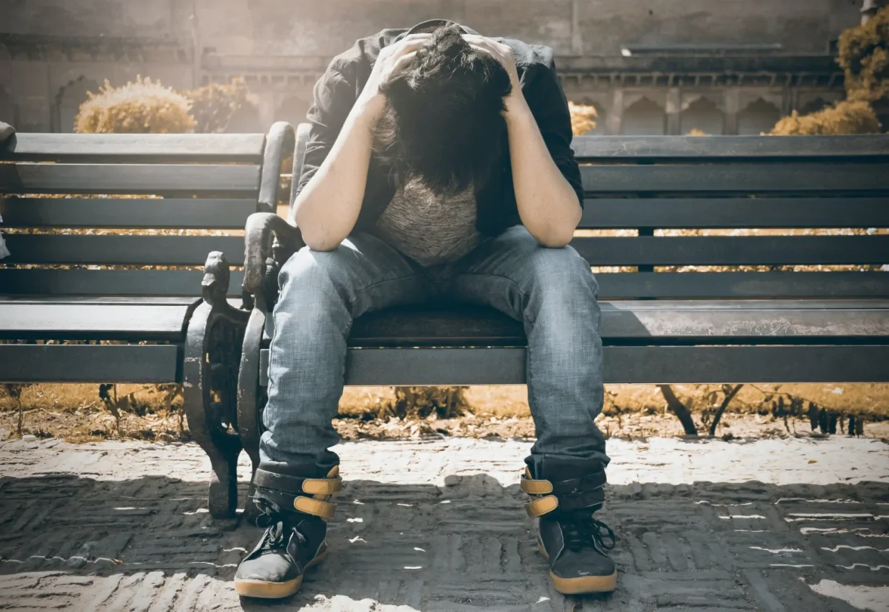 Jonge jongen op een houten bankje buiten die met zijn hoofd in zijn handen zit wegens verslaving.