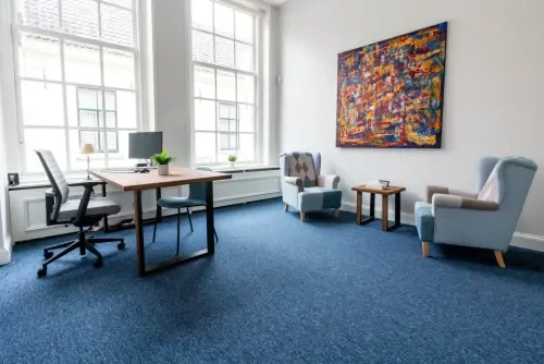 Behandelkamer met blauw tapijt, witte muren en een bureau met bureautstoel plus twee fauteuils.