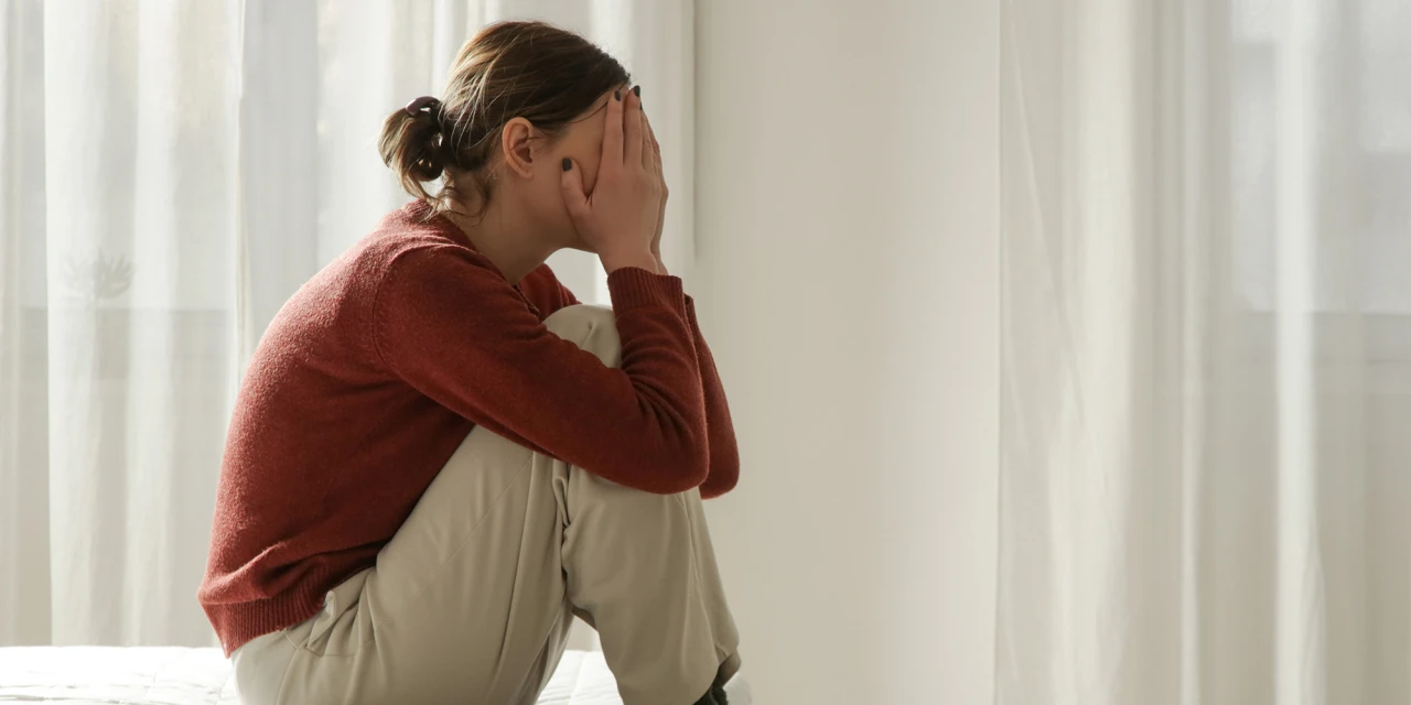Vrouw zit op bank met handen voor gezicht, zichtbaar overbelast door stress.