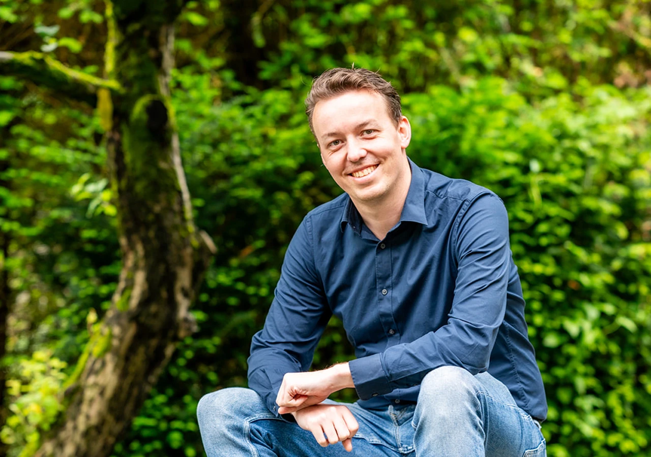 Lachende man in een donkerblauw overhemd en spijkerbroek zit voor een groene achtergrond van wazige bomen en planten.