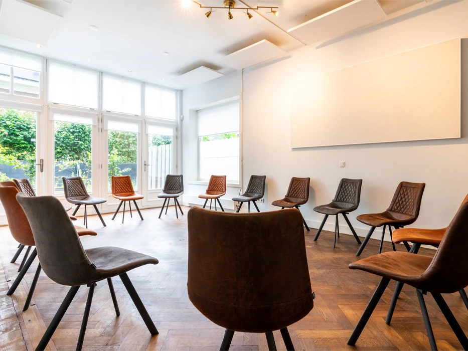 Lichte therapieruimte met visgraatvloer en stoeltjes in groepsopstelling en een presentatiebord. 