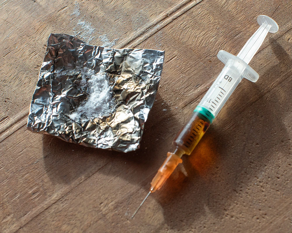 Een injectiespuit ligt naast wit poeder op aluminiumfolie op een houten ondergrond, passend bij crack- en harddrugsgebruik.
