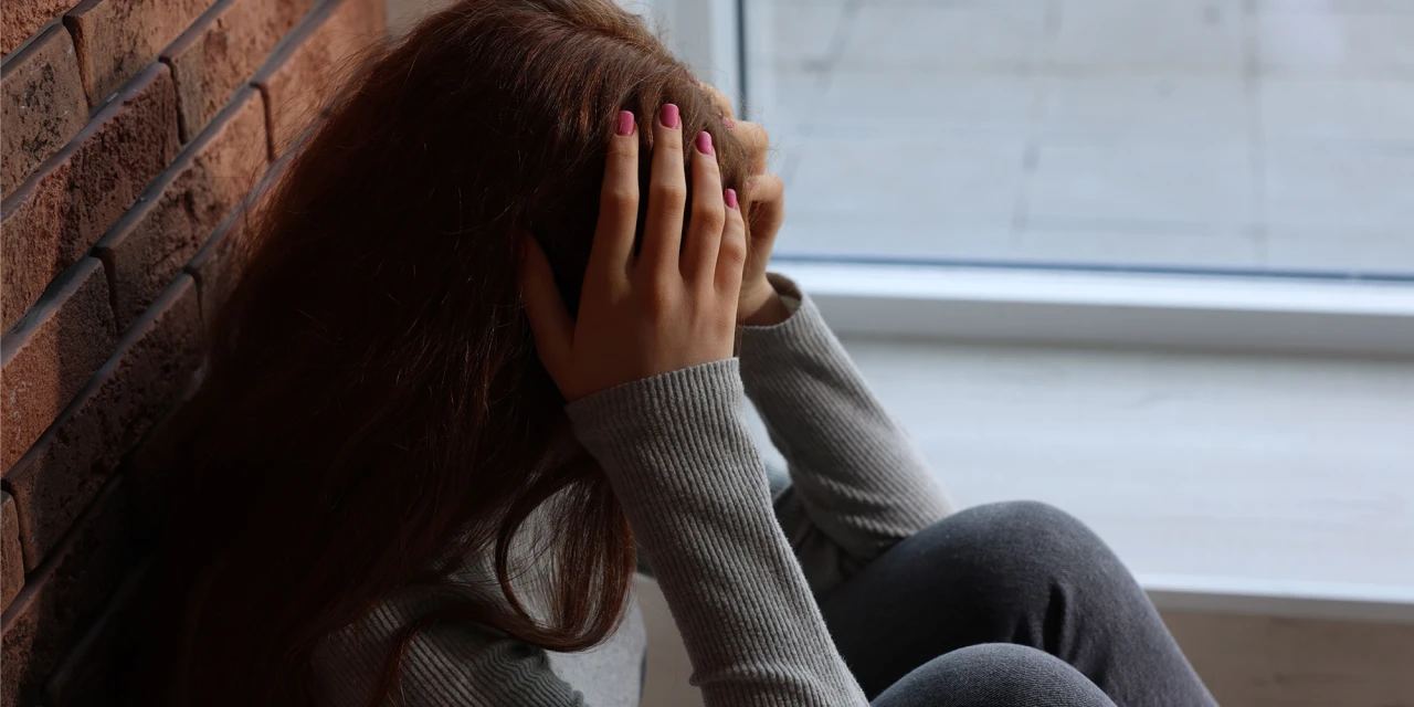 Persoon zit tegen muur met hoofd in handen bij raam tijdens dinsdagdip.