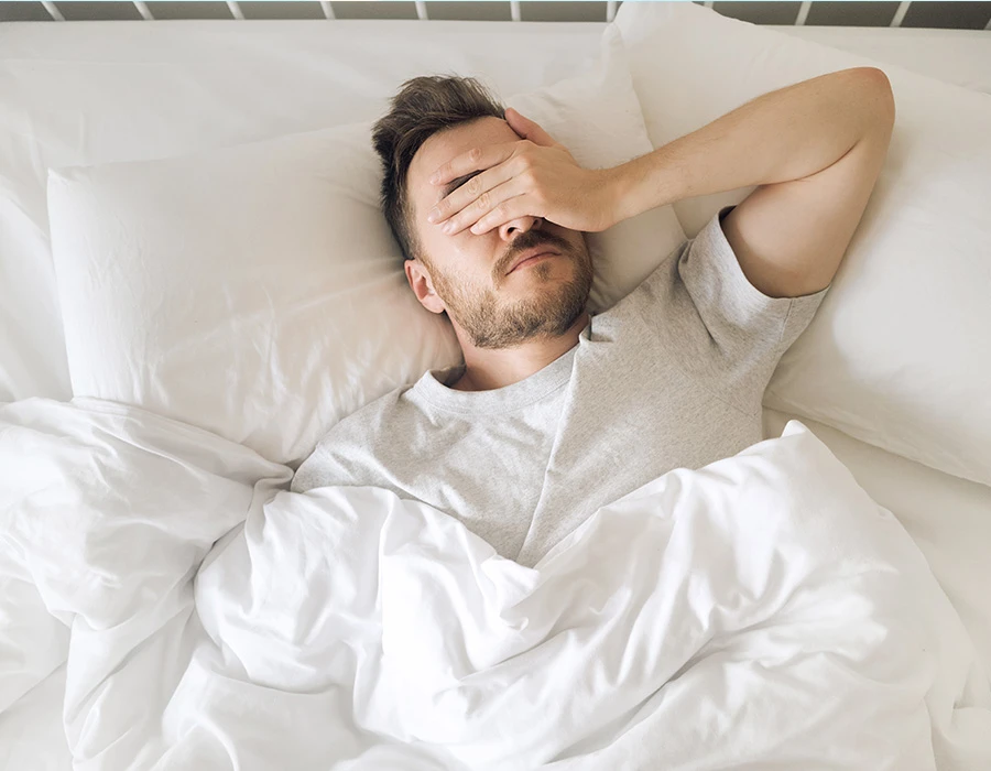 Man in bed houdt zijn hand voor zijn ogen tegen het licht, ter illustratie van een kater.