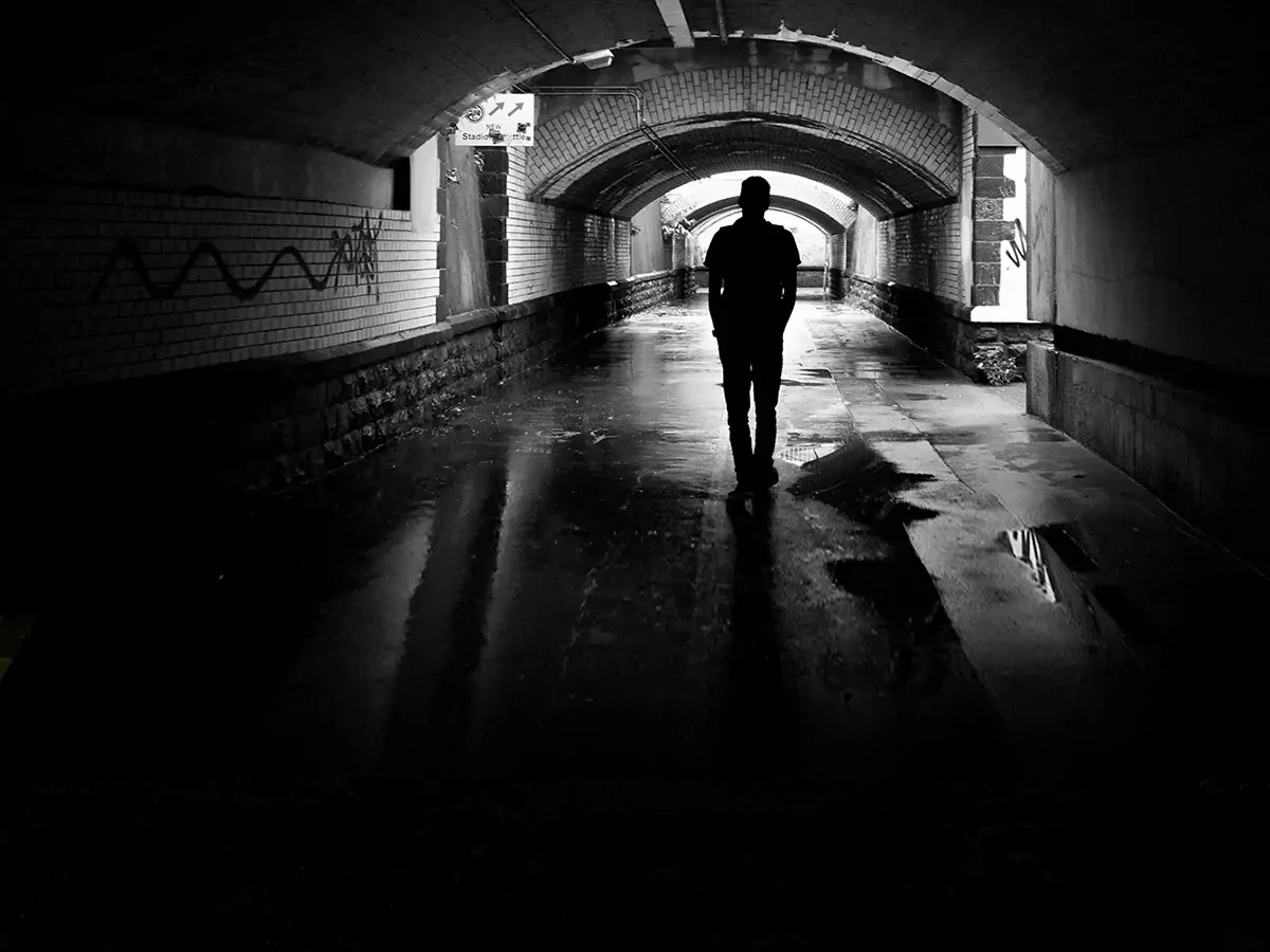 Zwart-witte foto met het silhouette van een man die door een donkere steeg loopt. 