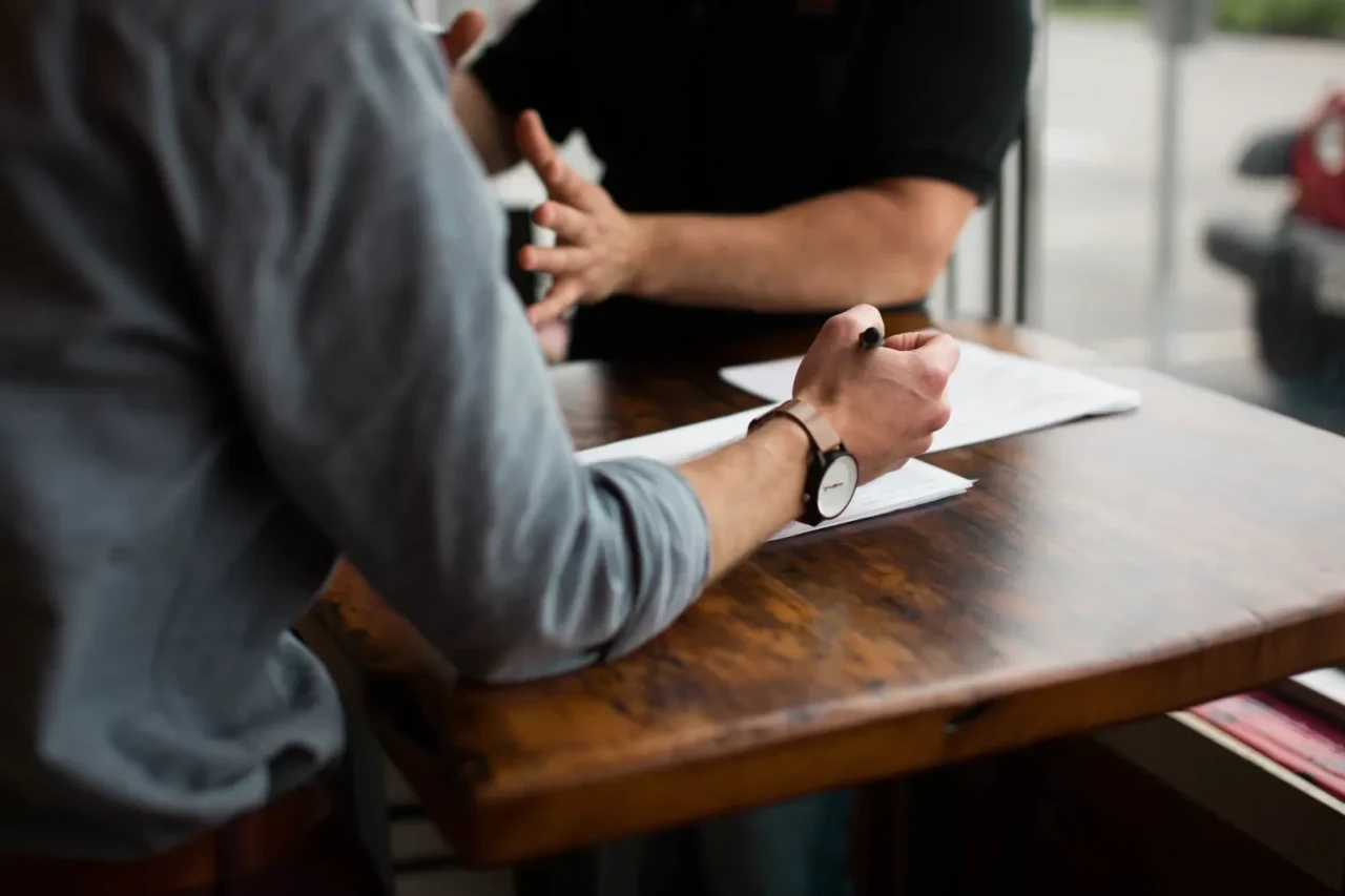 intakegesprek-aan-tafel Intakegesprek vindt plaats aan tafel tussen twee personen met vellen papier voor zich.