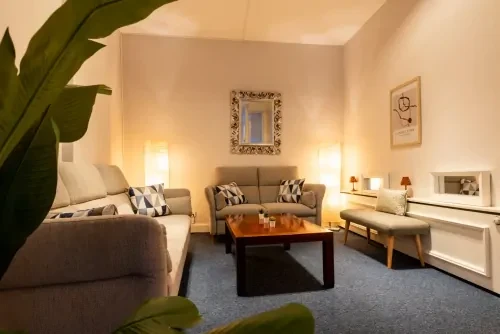 Warm verlichtte woonkamer met twee banken met kussens, witte muren en een rechthoekige salontafel.
