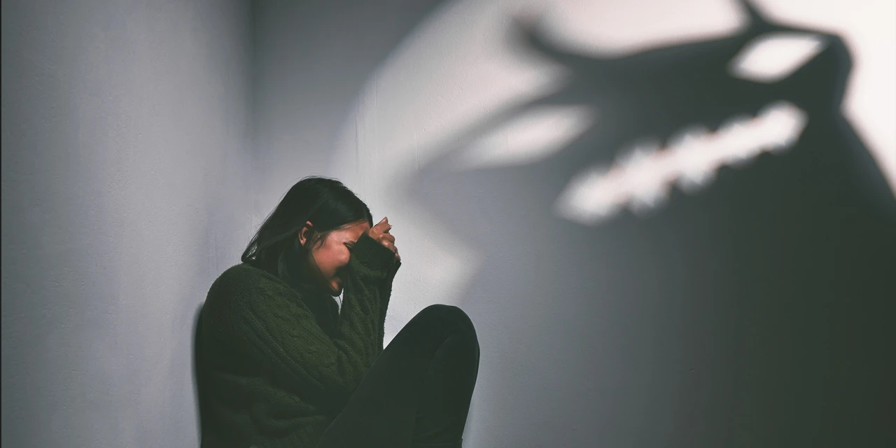 Persoon zit huilend in hoek, dreigende schaduw, gevoel van angst en isolatie.