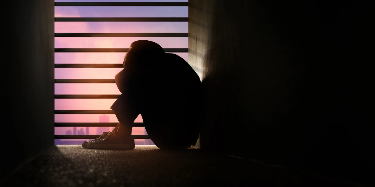 Persoon zit ineengedoken bij raam bij zonsondergang.