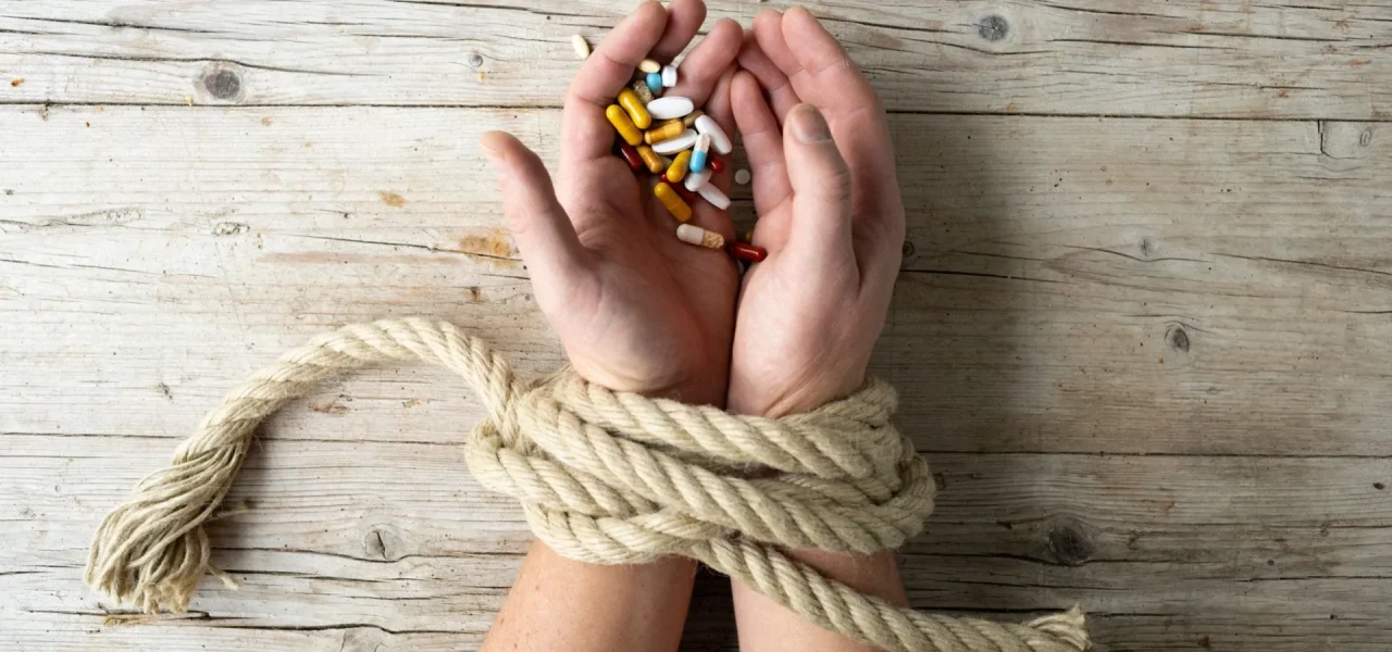 drugs-in-gebonden-handen Met touw aan elkaar gebonden handen die allerlei pillen vasthouden en op een houten tafel liggen.