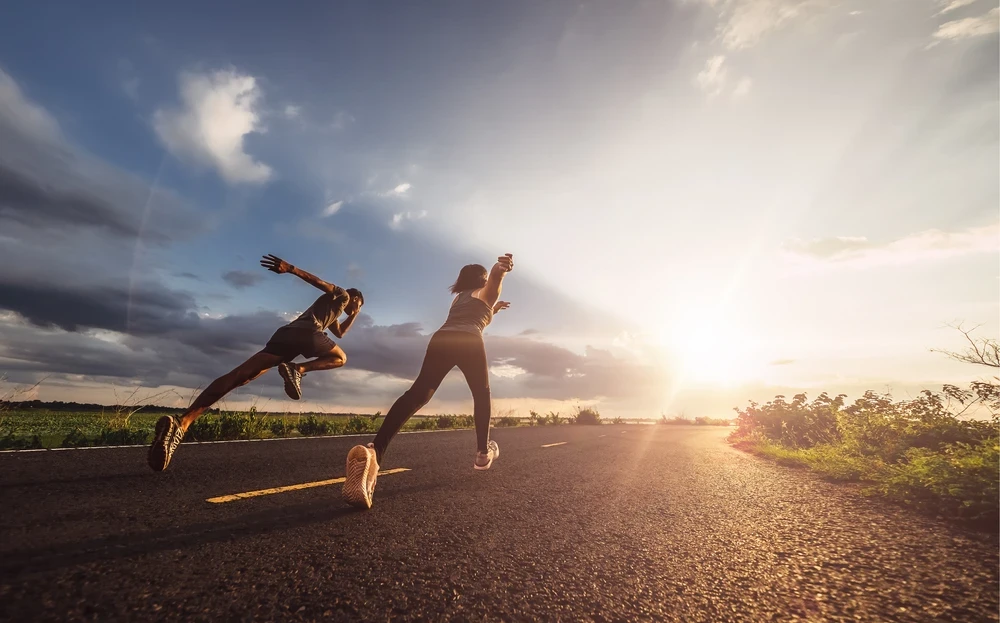 Twee sprintende mensen op een lege weg met een felle zon en blauwe lucht op de achtergrond.