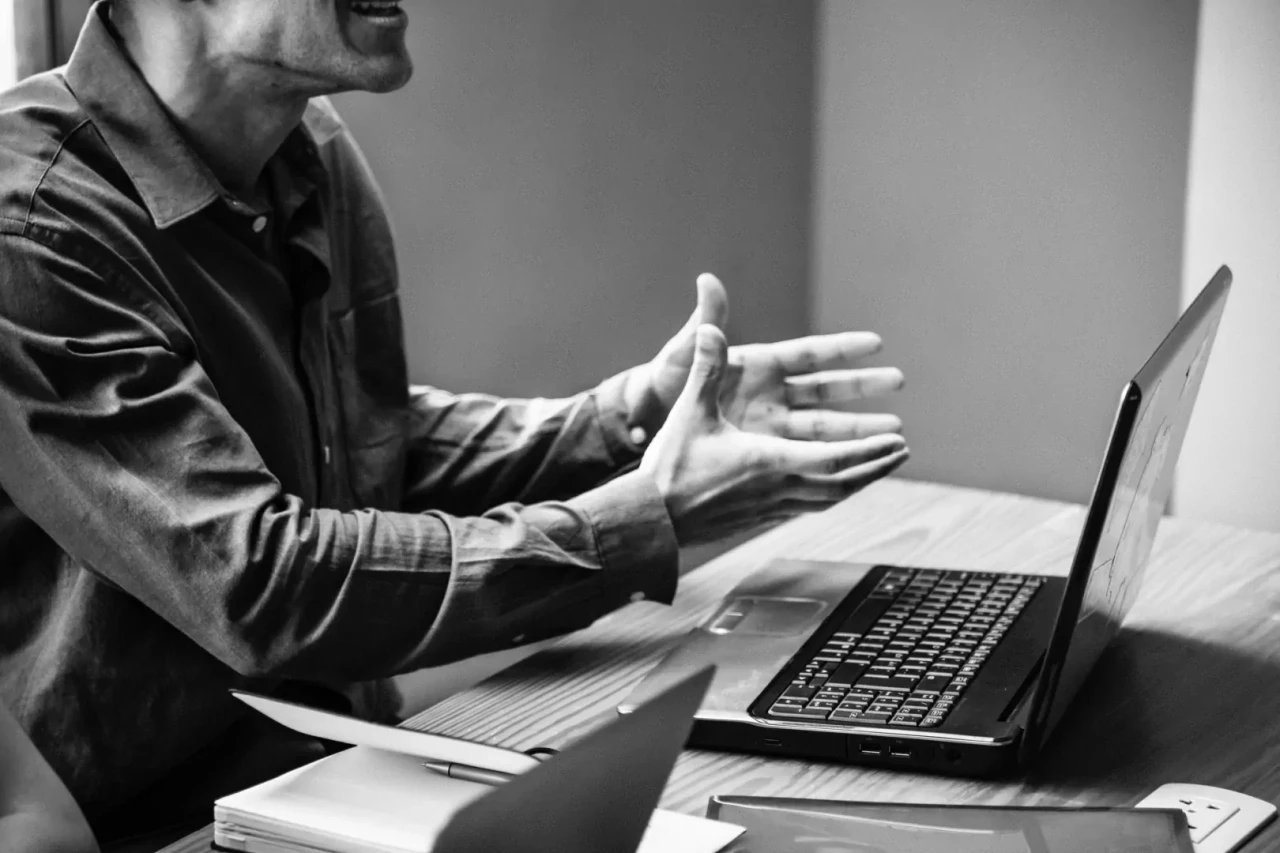 vragende-man-achter-laptop Zwart-wit foto van een man achter een tafel met laptop en die de handen vragend uitstrekt.
