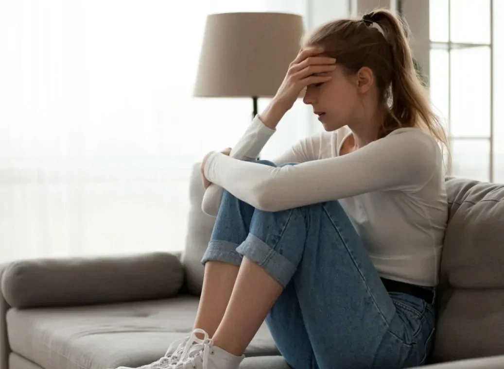 depressieve-vrouw-op-bank Jonge vrouw zit met de benen opgetrokken op de bank en houdt haar hand tegen haar voorhoofd.
