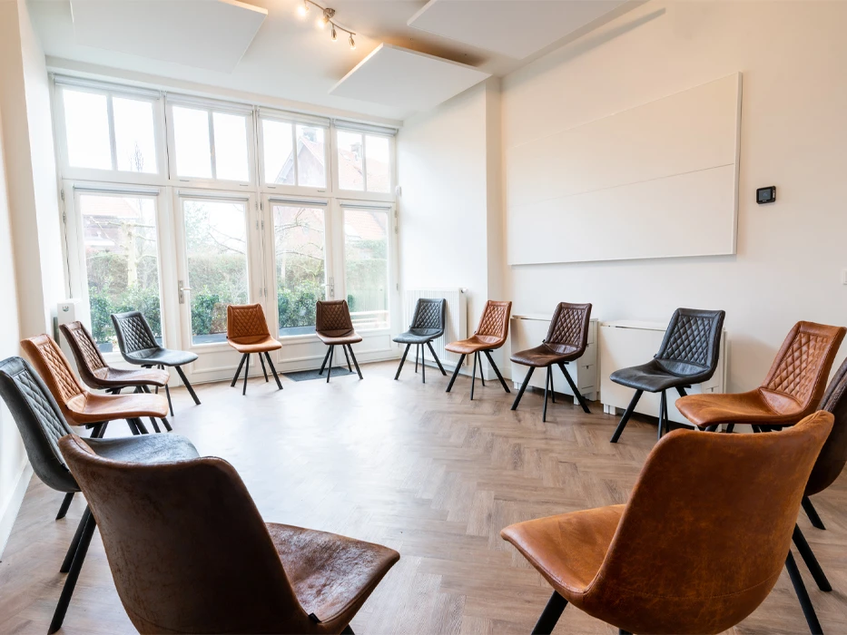 Behandelkamer met witte muren, een grote raampartij en 14 bruinleren stoelen in een opstelling voor kringgesprek.