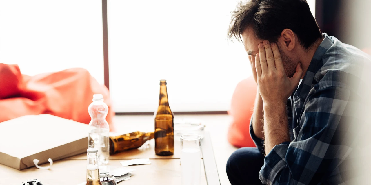 Man zit met handen voor gezicht tussen lege flessen en denkt wanneer ben je alcoholist?