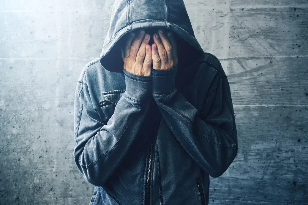 Staande, depressieve man in vest met capuchon op en de handen in het gezicht drukkend.