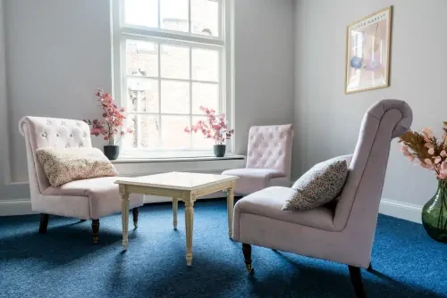 Behandelkamer met blauw tapijt, witte muren, drie lichte stoelen die tegen over elkaar staan met een salontafeltje in het midden.