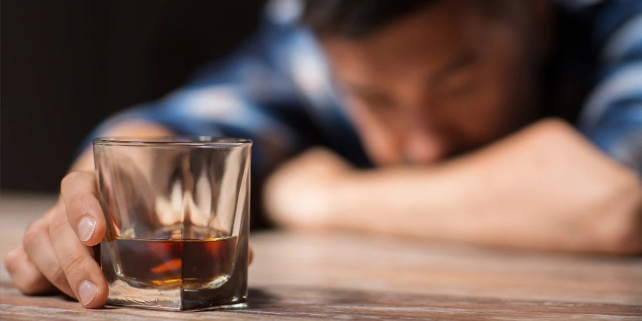 Man ligt met hoofd op tafel en houdt glas whisky vast na terugval alcohol.