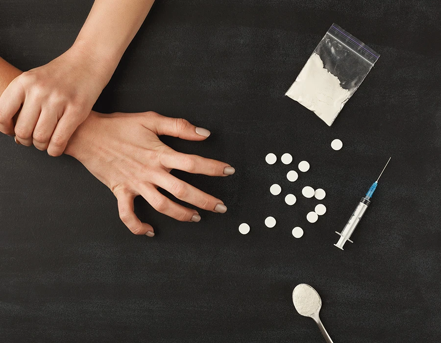 Handen naast drugs, pillen, een zakje poeder en een injectiespuit.