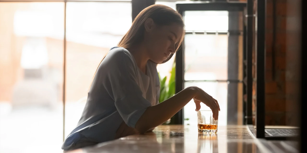 Alcoholverslaving bij vrouwen afgebeeld door vrouw die alleen met whisky aan tafel zit.