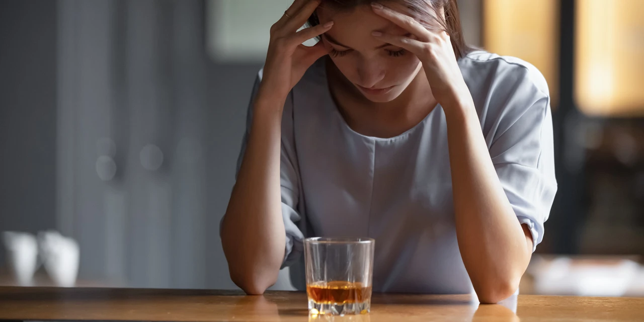 Vrouw zit bezorgd met glas alcohol aan tafel en toont uiterlijke symptomen alcoholist.